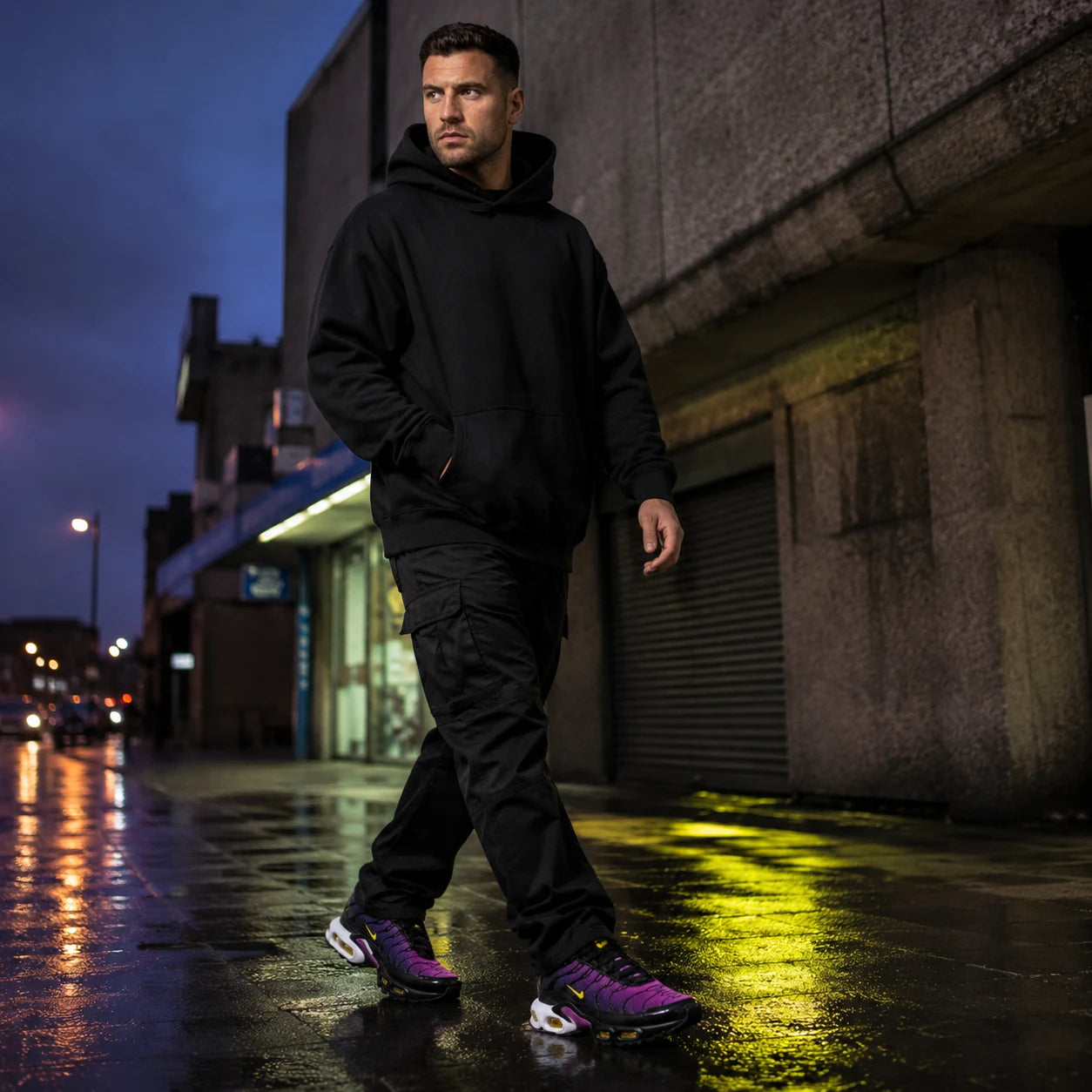 Urban Kinetic Wear lifestyle campaign — UK streetwear figure in black hoodie and cargos on a wet Tottenham street at dusk, wearing the TN Voltage Purple trainer with neon volt reflections on the pavement